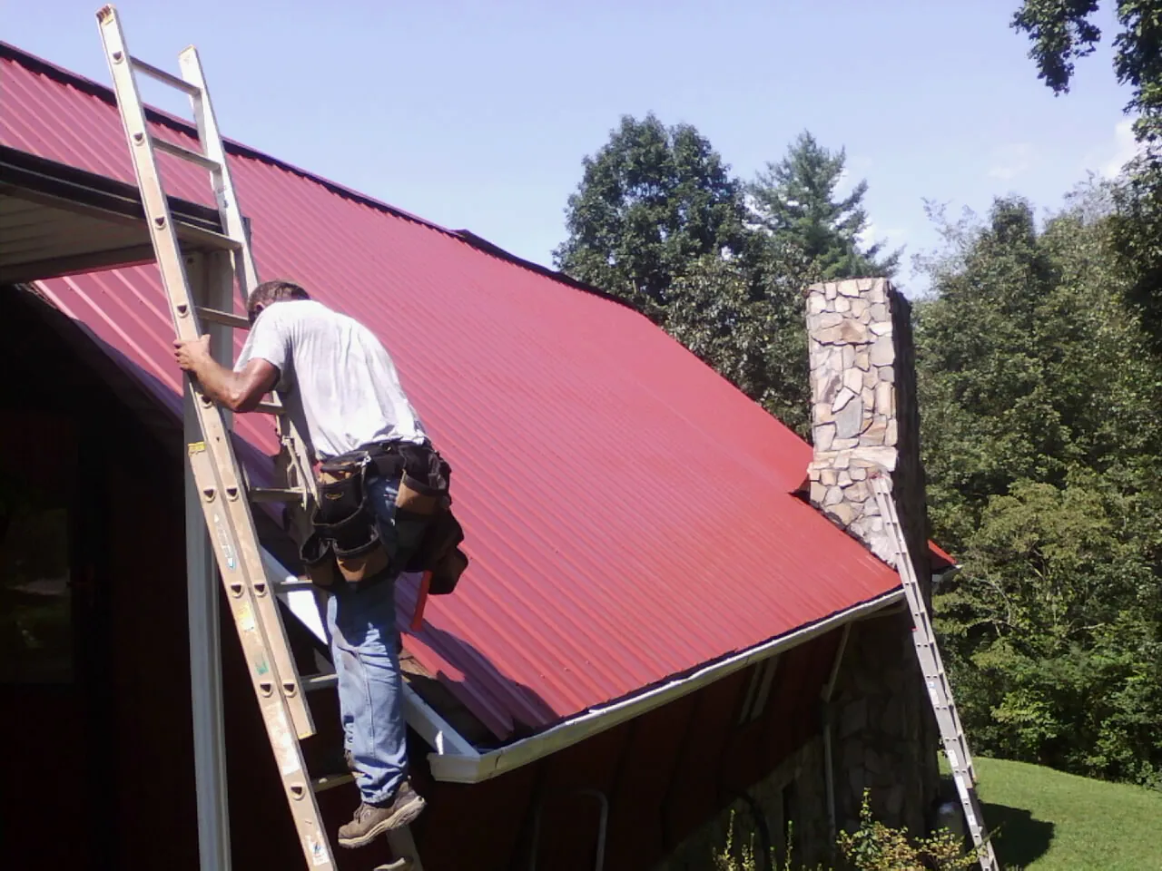 Roofer on a ladder beside a red metal roof and stone chimney — Soffit & Fascia Repair by Roofing Brothers in Shoemakersville